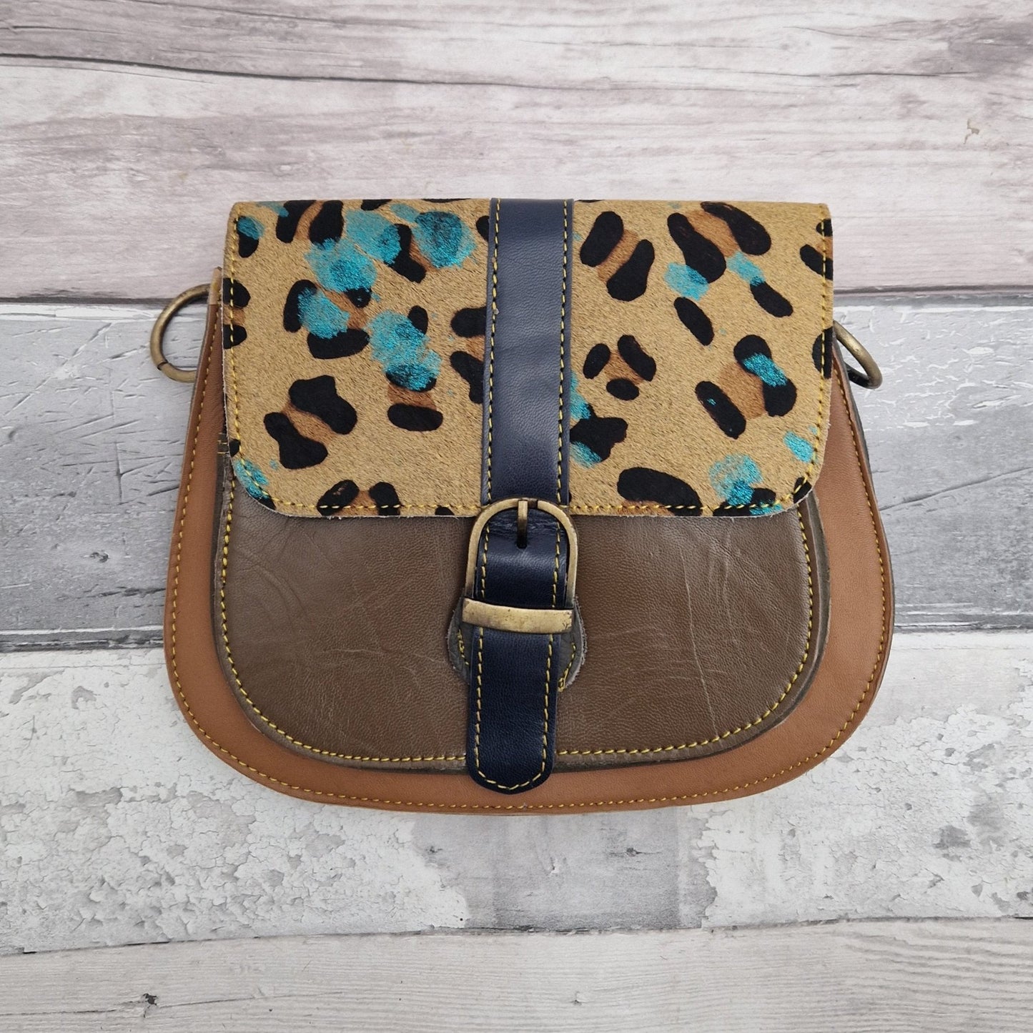 All leather saddlebag style ladies bag in 2 tones of brown and with a textured panel of leopard print splattered with metallic blue splashes. Brass buckle closer.