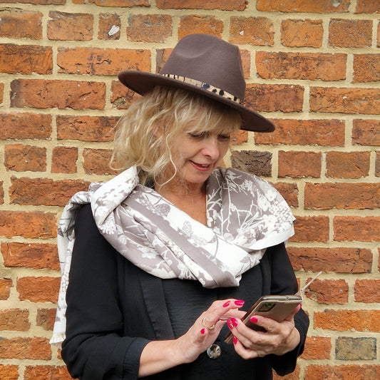 Lady wearing a Chocolate Brown Fedora Hat with a belted animal print band. She is wrapped in a cream and mocha coloured scarf with a Hedgerow scene print.