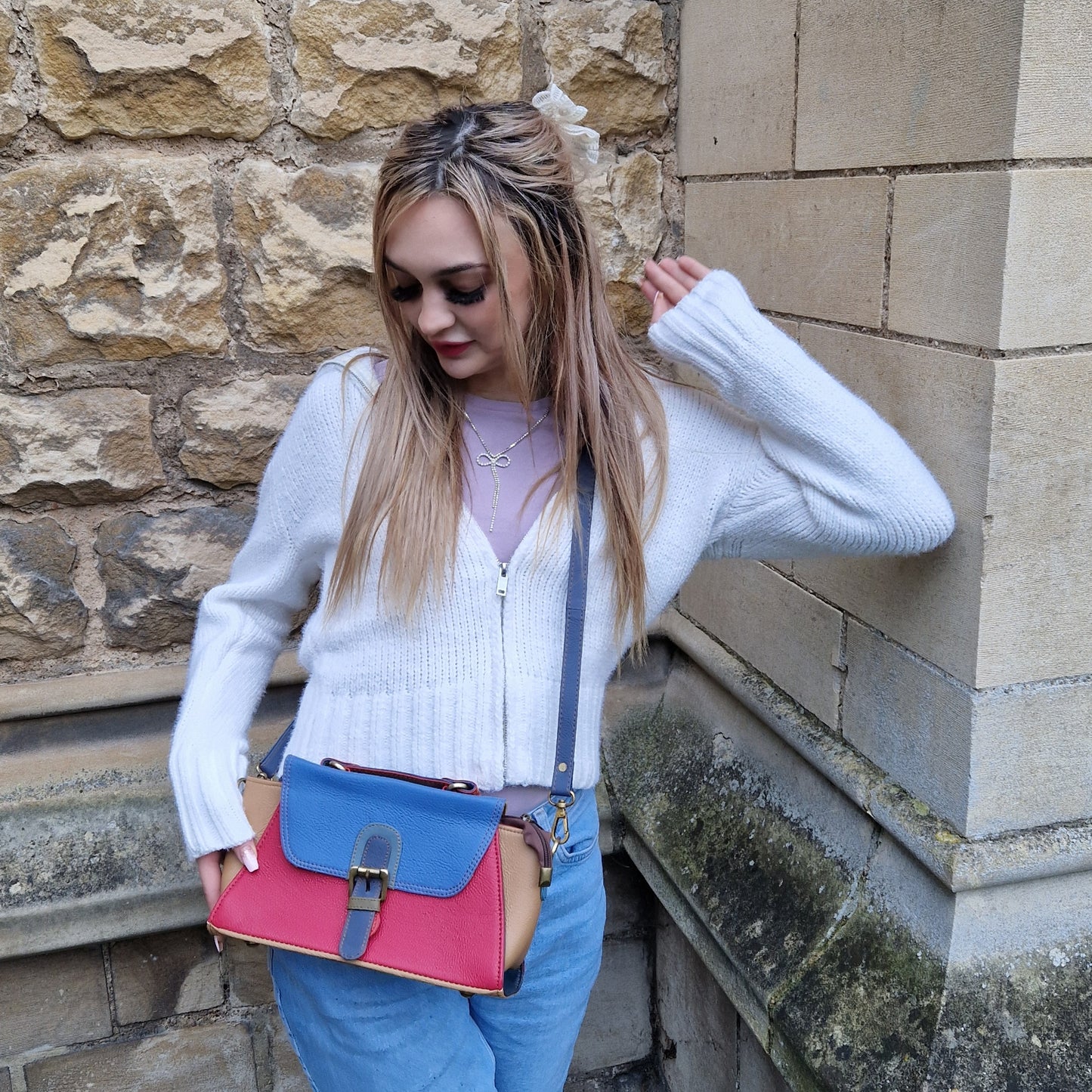 Young woman wearing a satchel style handbag across her body. Bag is brightly coloured and finished with a buckle front in a satchel style.