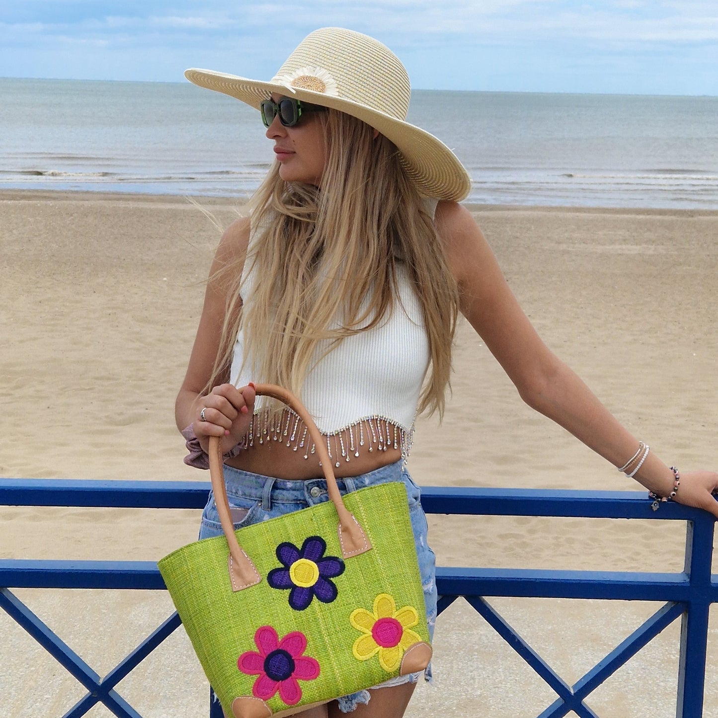 Young woman at the beach wearing a wide brim natural coloured sun hat, carrying a lime green raffia basket, decorated with flowers.