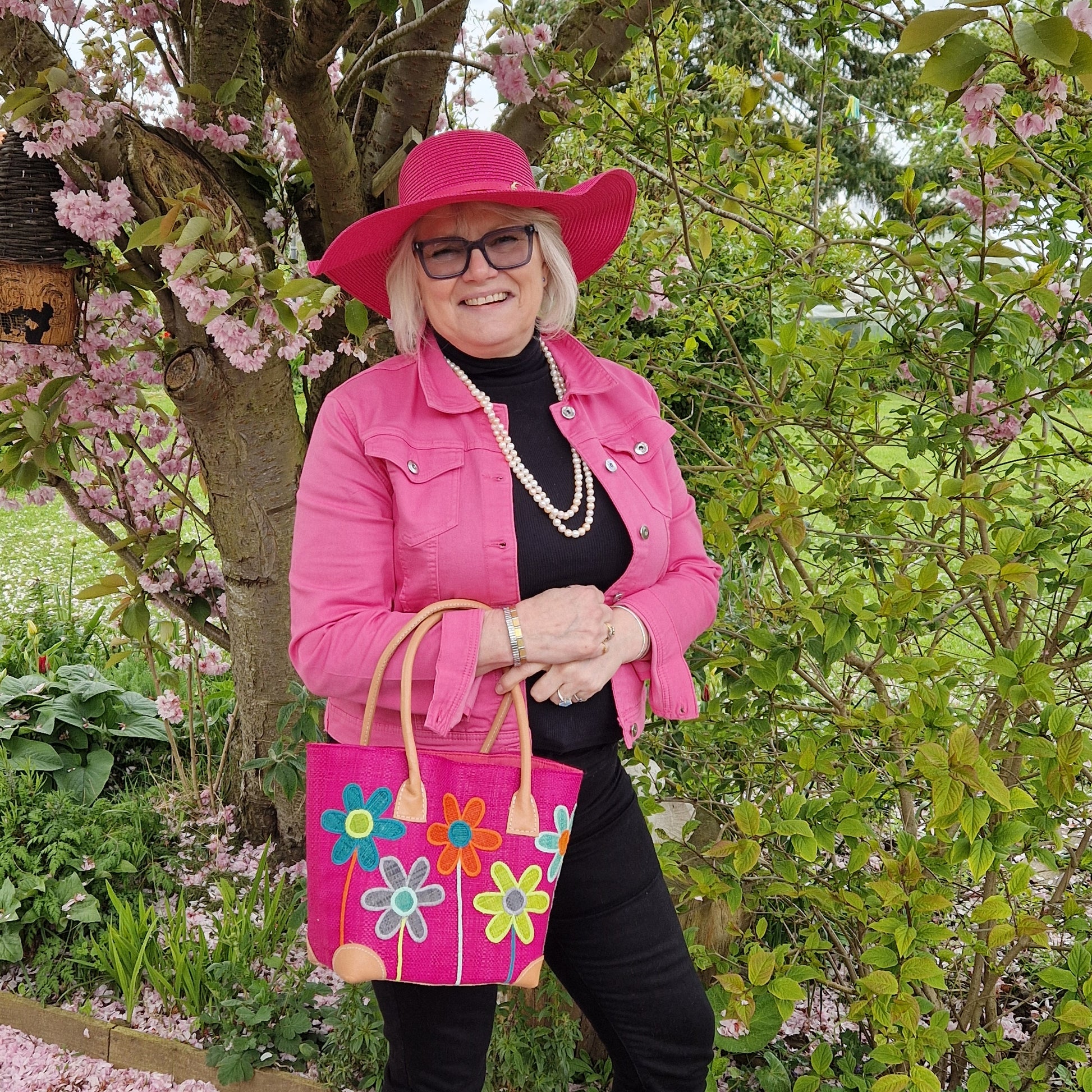 Lady wearing a wide brimmed hot pink sun hat and carrying a matching hot pink coloured raffia basket, decorated with brightly coloured flowers.