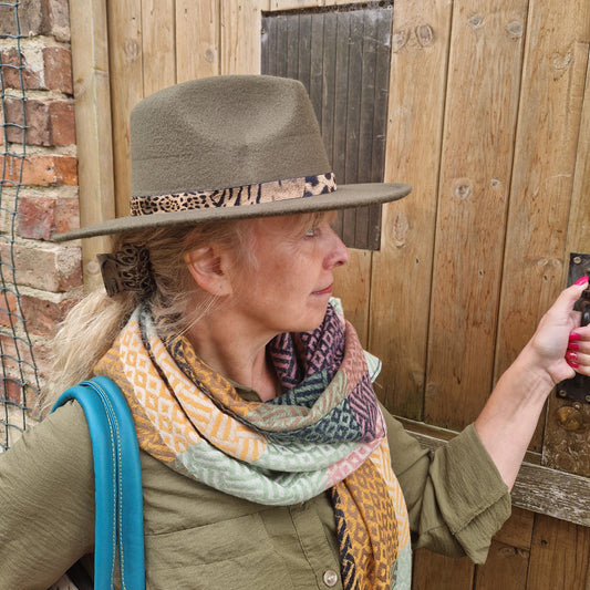 Woman wearing a green felt Fedora hat with an animal print belted band and a patchwork scarf in green with multi-coloured panels.