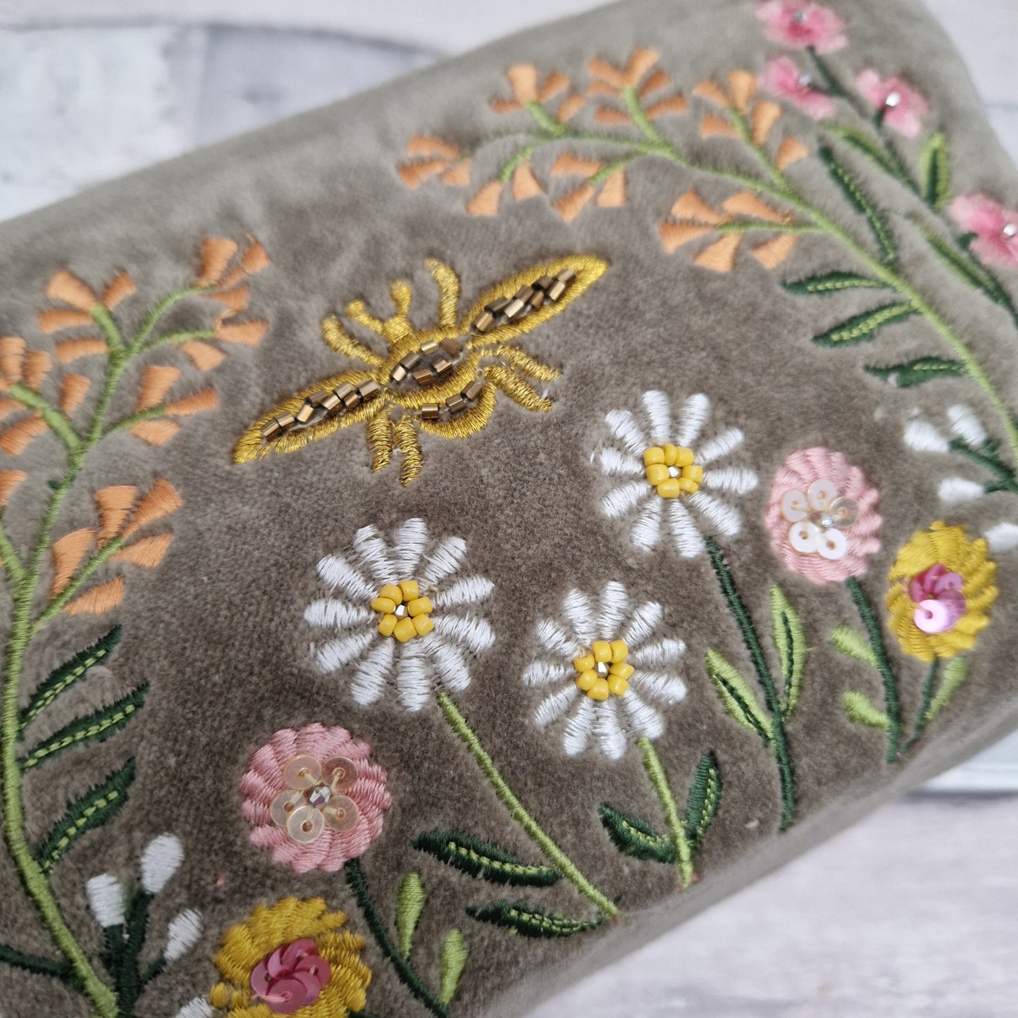 Close up view of our Sage coloured velvet washbag hand embroidered with sequins and beads. Depicting a flower garden with a central golden coloured bee.