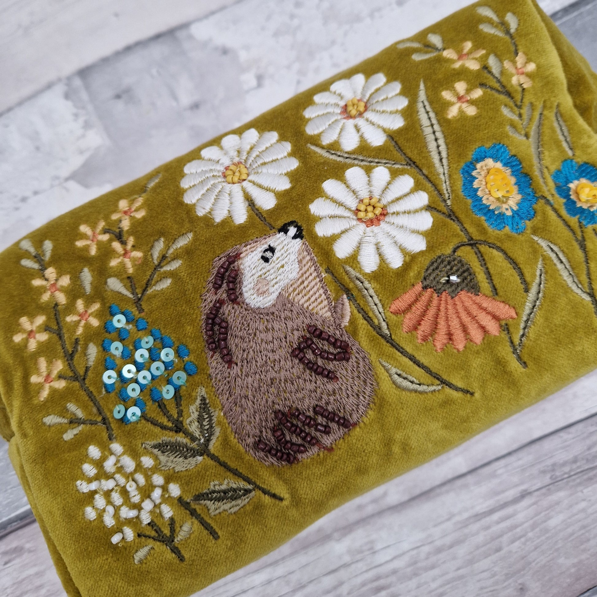 A Mustard colored velvet wash bag. Decorated with hand embroidery, sequins and beads, depicting a hedgehog in a wild flower meadow.