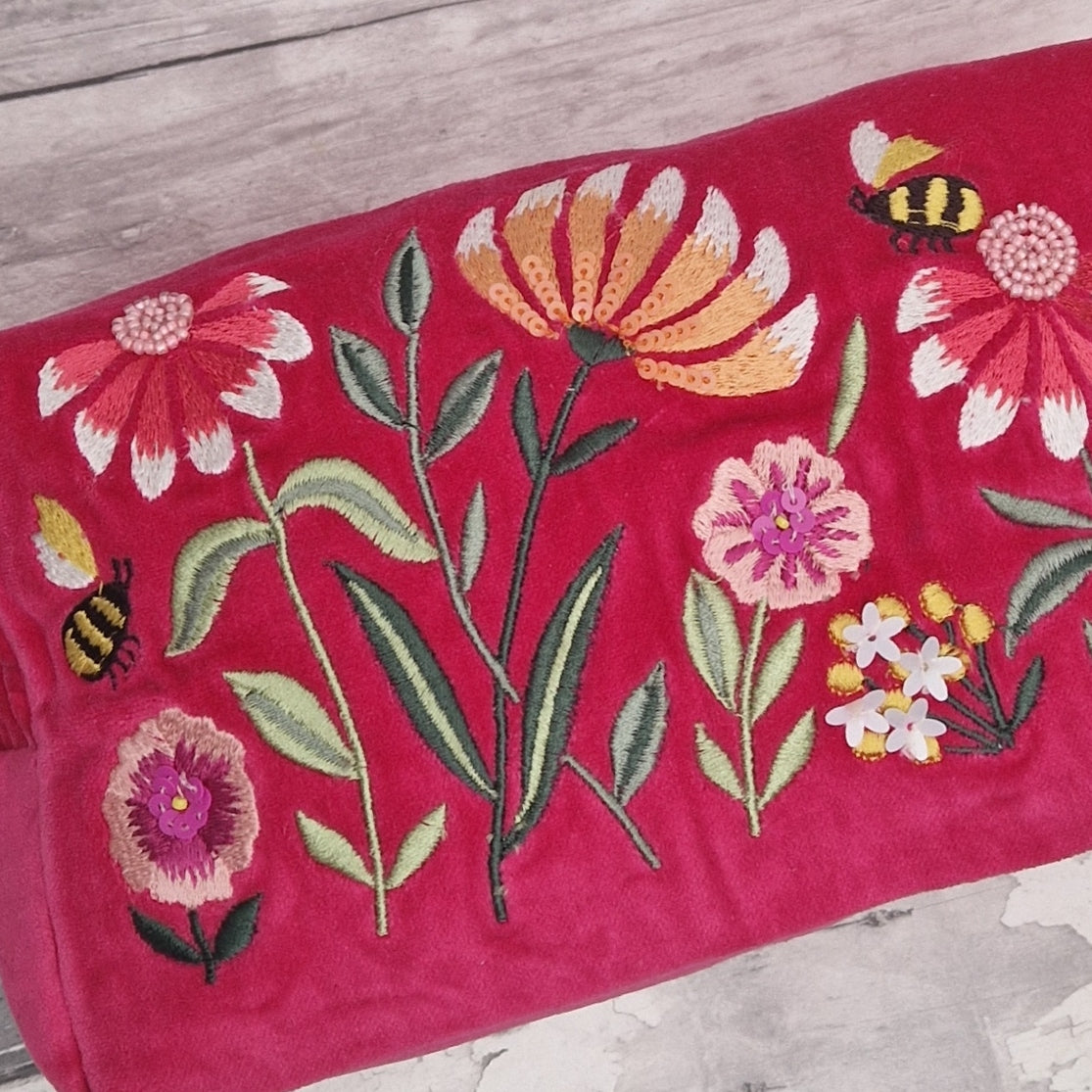 Close up view of a hot pink velvet washbag. Decorated with embroidery, hand sewn sequins and beads. Depicting a flower garden with bumble bees. 